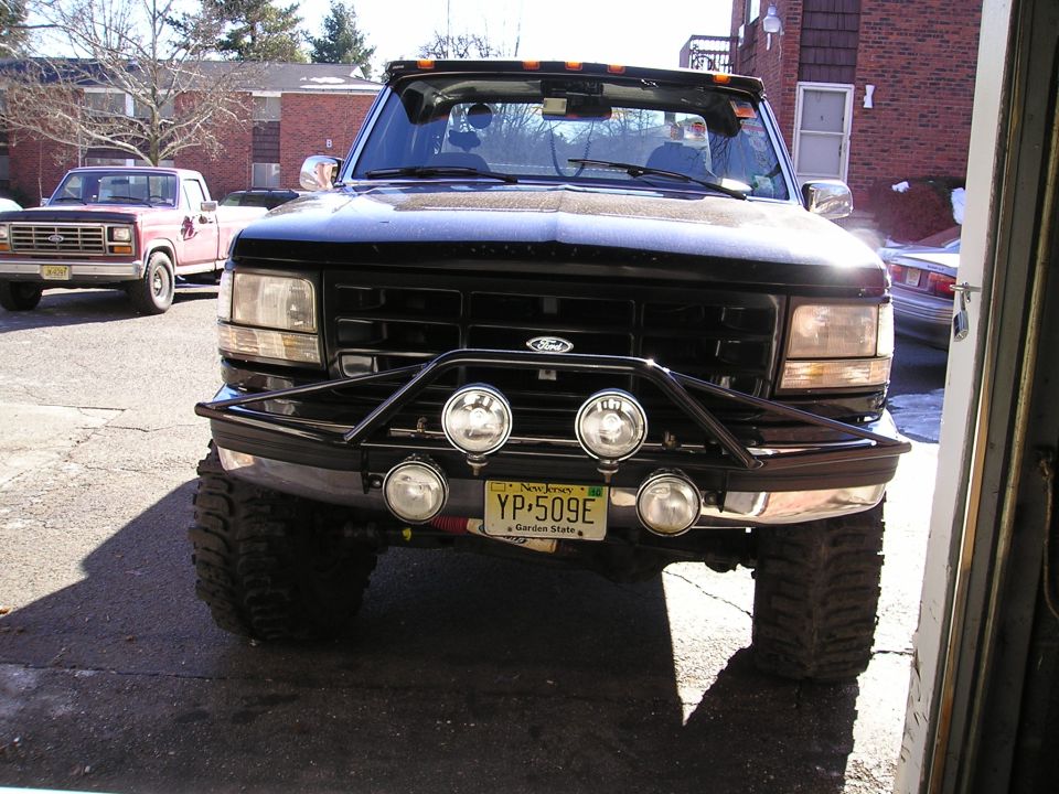 Front view of black Ford F-Series truck with a brush guard and four lights. Another red truck is parked behind it