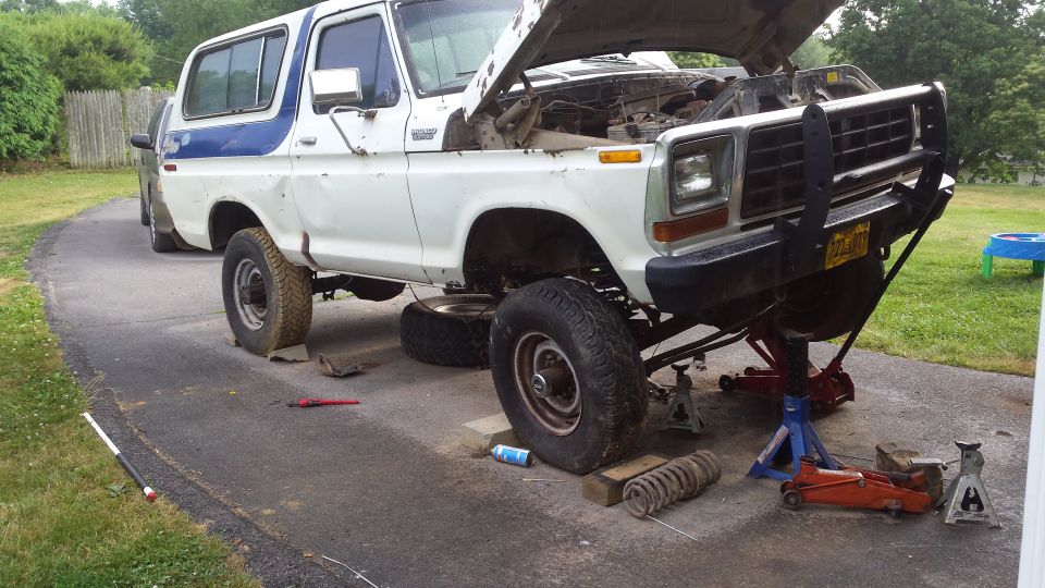 White 1979 Ford Bronco on jack stands, hood up, with coil spring removed.