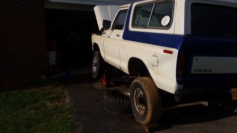 White and blue Ford Bronco with hood up, being worked on, one tire removed.