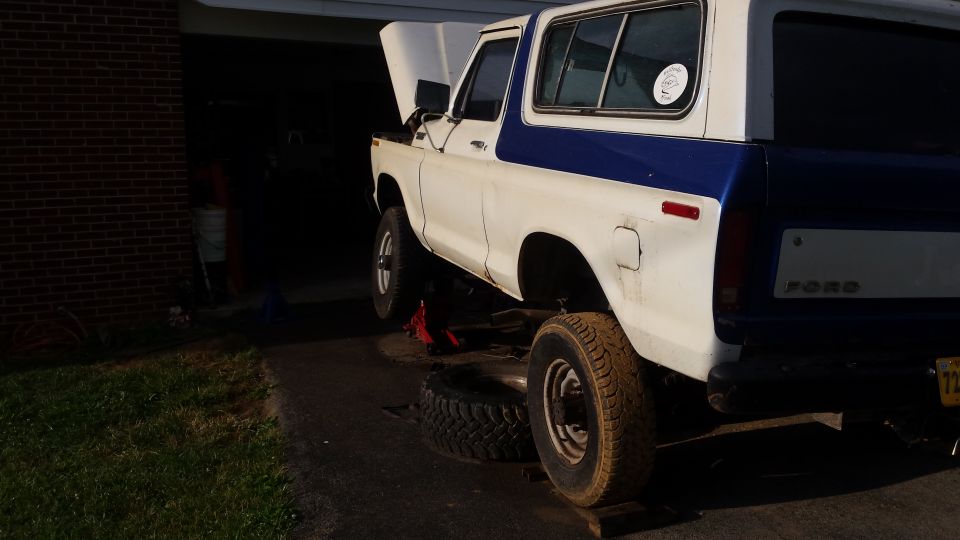 White and blue Ford Bronco with hood up, being worked on, one tire removed.