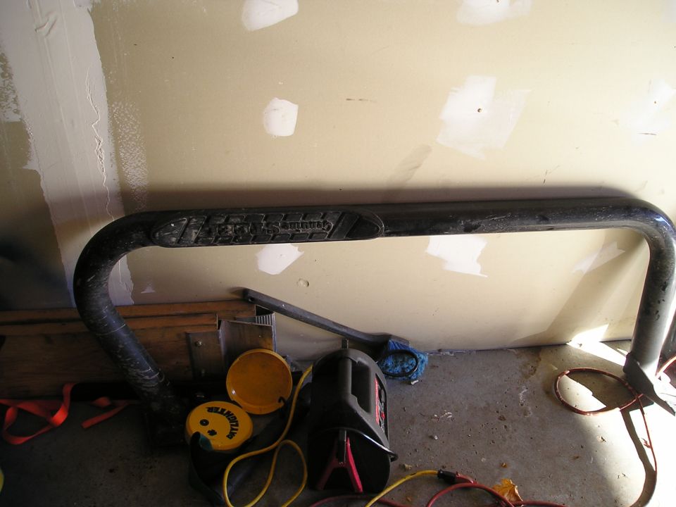 Black truck side step with textured grip area, leaning against a wall. A jump starter and yellow containers are on the floor