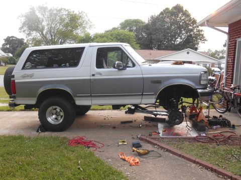 1994 Ford Bronco - A two-tone white and gray 1994 Ford Bronco is parked in a driveway with its front end raised on jack stands, undergoing maintenance. Variou…