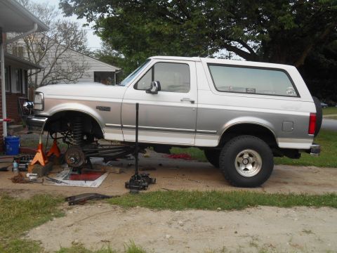 1994 Ford Bronco - A two-tone silver and white Ford Bronco is raised on jack stands with its front suspension disassembled. Various tools and parts are scatte…