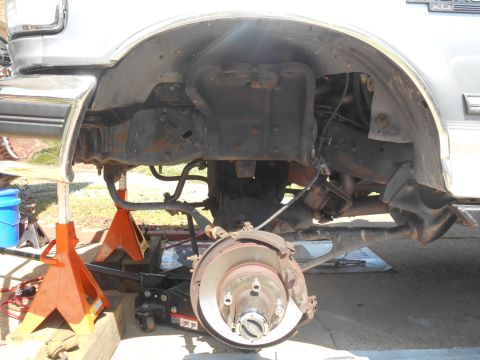 1994 Ford Bronco - The front suspension and wheel hub assembly of a silver Ford Bronco are visible, supported by jack stands and a floor jack. The vehicle app…