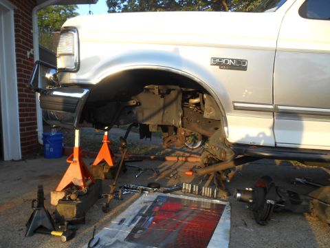 1994 Ford Bronco - The front end of a white Ford Bronco is raised on jack stands with the front wheel and suspension components removed and laid out on the gr…