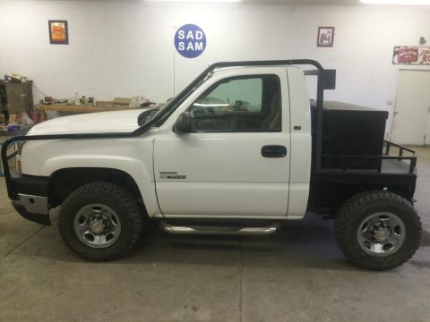 1985 Jeep CJ7 - A white, four-door pickup truck with a flatbed and a black toolbox is parked indoors on a concrete floor. The truck has large tires, a blac…