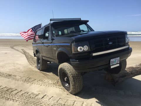 1992 Ford Bronco - A black 1992 Ford Bronco is parked on a sandy beach with an American flag attached to the driver's side. The vehicle features aftermarket h…