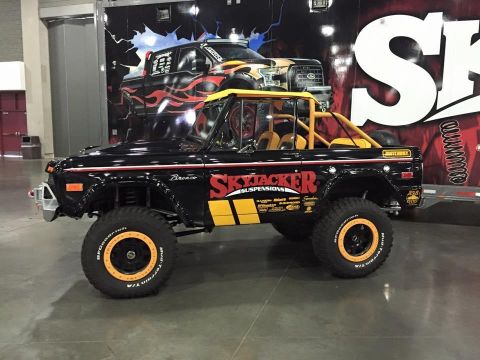 1982 Ford F-100 - A black 1970s Ford Bronco with yellow accents and large off-road tires is parked indoors in front of a trailer with a graphic of a Ford F-1…