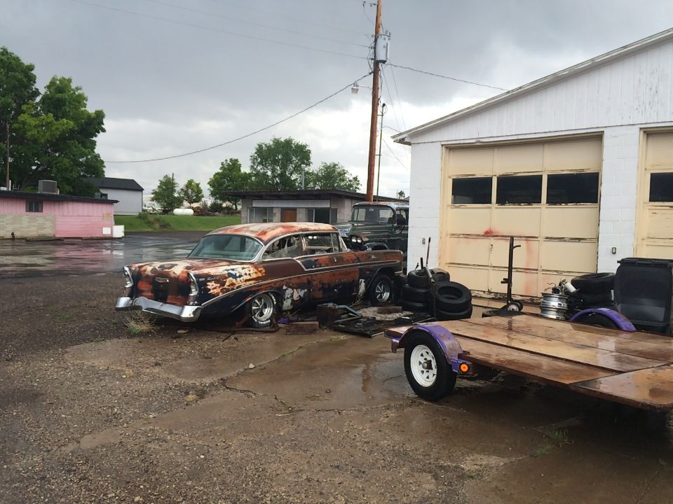 Photo of a rusty project car and trailer in a lot by a garage
