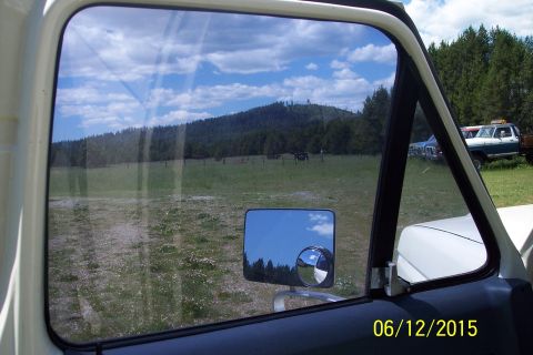 1990 Ford Bronco - View through the driver's side window of a vintage Ford truck, reflecting a cloudy blue sky and a tree-covered hill. A side mirror with a c…
