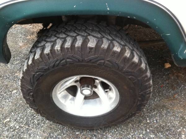Close-up of a knobby off-road tire on a silver five-spoke wheel, mounted on a dark green SUV.