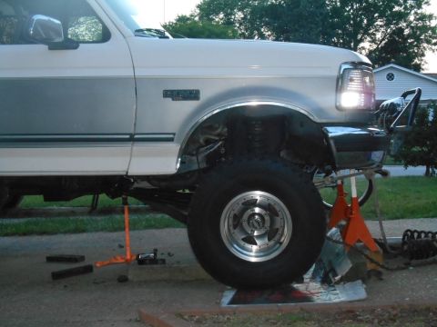 1994 Ford Bronco - The driver's side of a white 1994 Ford Bronco is shown lifted on jack stands with the front wheel removed. The vehicle appears to be underg…