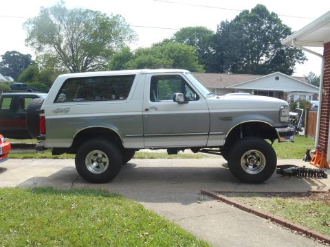 1994 Ford Bronco - A lifted two-tone white and gray 1994 Ford Bronco is parked on a paved driveway in front of a house. The vehicle has aftermarket wheels and…