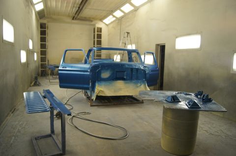 1979 Ford Bronco - A blue Ford truck cab is being painted in a spray booth. Various parts of the truck and painting equipment are visible around the vehicle.
