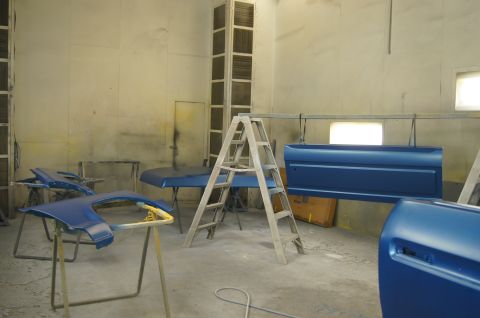 1979 Ford Bronco - A car paint booth contains several blue car body parts, including a fender, hood, and doors, resting on stands and hanging from a rack.