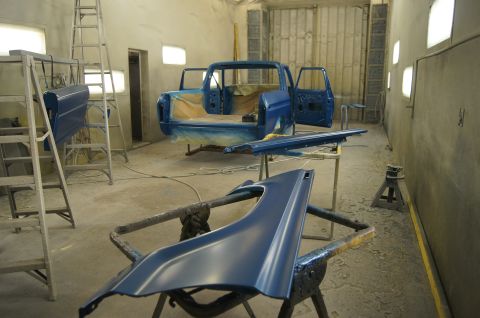 1979 Ford Bronco - A blue pickup truck body and several parts are in a paint booth, with ladders and stands supporting them. The truck bed and cab are visible…