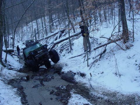 1992 Ford Bronco - An off-road vehicle navigates a difficult path through a snowy, wooded area with fallen trees.