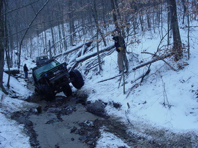 Jeep struggling on a muddy, snowy trail with fallen trees. Another Jeep is visible behind it