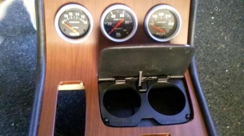 1982 Ford Bronco - A close-up view of a wooden dashboard panel with three gauges and a flip-top cup holder. The gauges measure voltage, oil pressure, and wate…