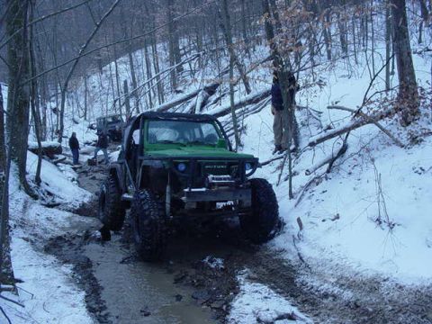 1992 Ford Bronco - Multiple off-road vehicles navigate a challenging muddy and snowy path through a wooded area.
