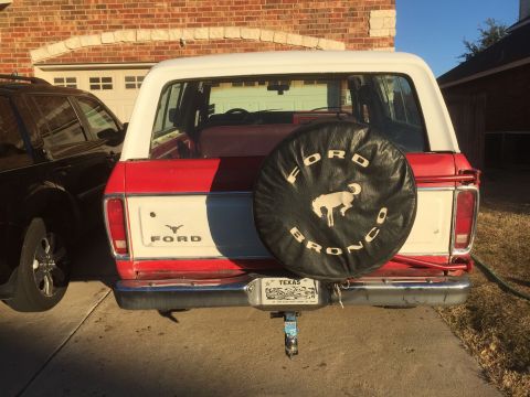 1979 Ford Bronco - The rear of a red and white 1979 Ford Bronco is shown parked in a driveway next to a black SUV. A spare tire cover with a horse graphic and…
