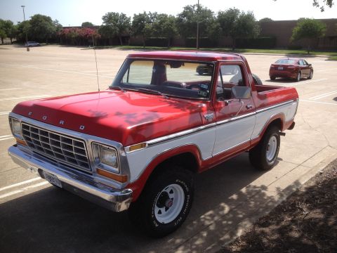 1979 Ford Bronco - A red and white 1979 Ford Bronco is parked in a paved lot with a red sedan in the background. The Bronco has a chrome grille and bumper, an…