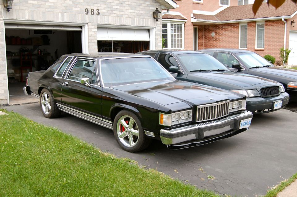 Black 1985 Mercury Grand Marquis with chrome trim and custom wheels parked in a driveway.