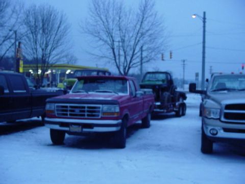 1992 Ford Bronco - A red pickup truck is parked in a snowy lot next to other vehicles, including one on a trailer.