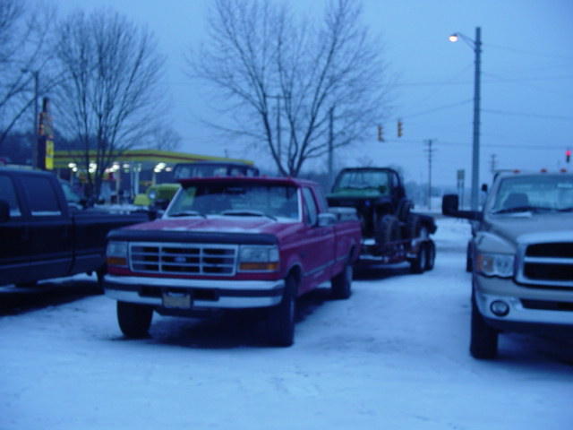Red pickup truck with a dark grille and headlights, parked in snow. Another truck is on a trailer behind it