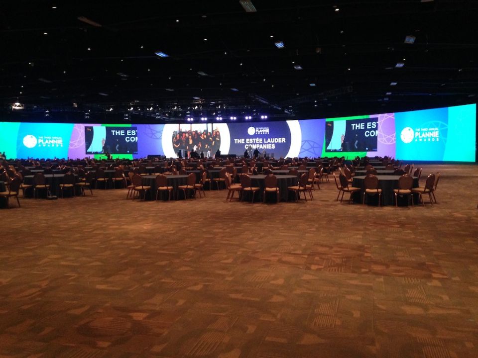 Conference hall with round tables and chairs facing a large LED screen displaying event graphics.