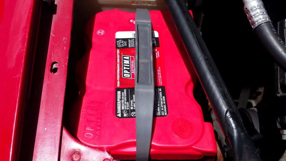Red Optima battery in a red vehicle compartment with a black strap.