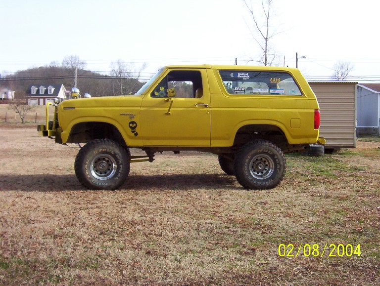 Yellow lifted Ford Bronco on mud-terrain tires parked on grass beside a metal shed