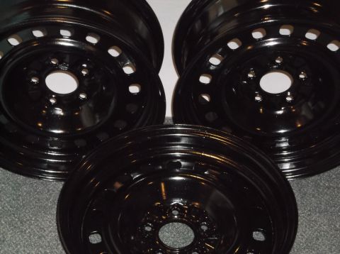 1999 Mercury Grand Marquis - Three black steel wheels are arranged on a gray carpet. The wheels have multiple circular holes and a central hub opening.