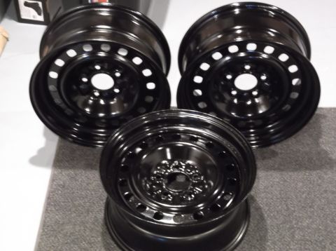 1999 Mercury Grand Marquis - Three black steel wheels are arranged on a gray carpeted surface. The wheels have multiple circular holes and a central hub opening.