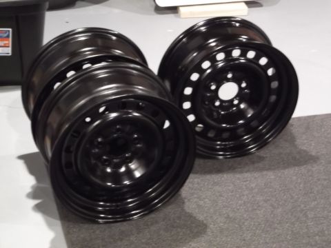 1999 Mercury Grand Marquis - Two black steel wheels are stacked and leaning against each other on a gray carpeted surface. The wheel on the right is fully visible, show…
