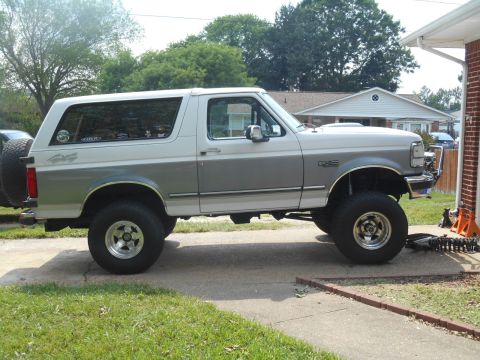 1994 Ford Bronco - The side profile of a lifted two-tone white and gray Ford Bronco is shown parked on a concrete driveway. A spare tire is mounted on the rea…