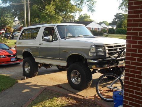 1994 Ford Bronco - A lifted, two-tone white and grey Ford Bronco is parked on a driveway next to a red Ford Mustang. The Bronco has a custom front bumper with…