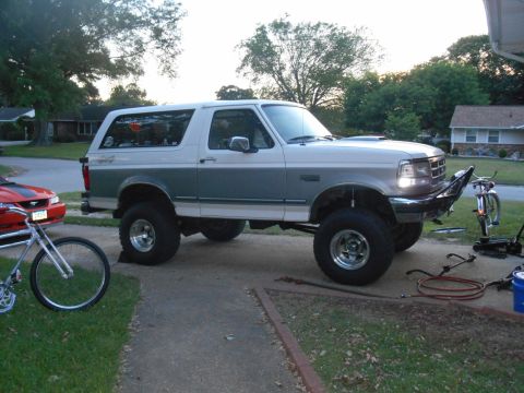 1994 Ford Bronco - A lifted two-tone white and teal Ford Bronco is parked on a driveway next to a red Mustang. A bicycle is parked in front of the Bronco.