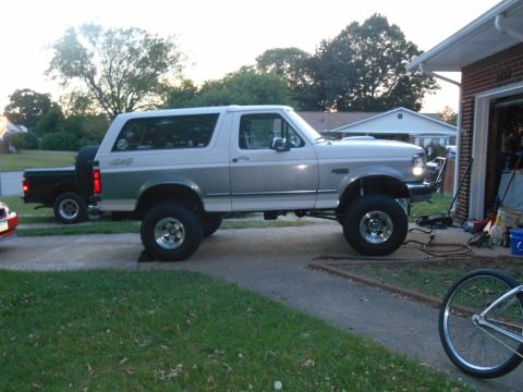 1994 Ford Bronco - A lifted, two-tone (white and silver) Ford Bronco is parked on a driveway next to a brick garage. A black pickup truck is parked behind it.
