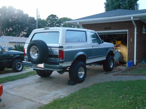 1994 Ford Bronco - The rear driver's side of a lifted, two-tone white and blue Ford Bronco is parked on a concrete driveway next to a brick garage. A spare ti…