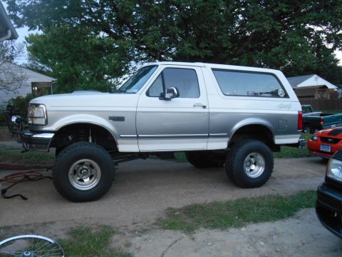 1994 Ford Bronco - A lifted, two-tone white and silver 1994 Ford Bronco is parked on a gravel driveway next to a house. The SUV has large off-road tires and a…