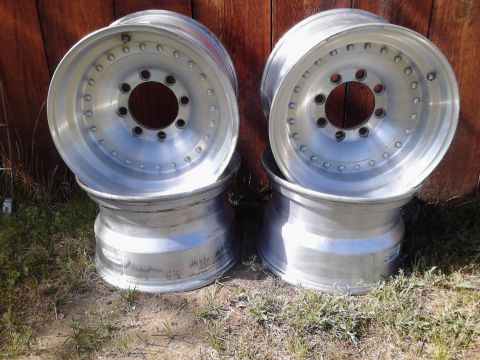 1989 Ford Bronco - A set of four aluminum alloy wheels are stacked and displayed outdoors against a wooden fence.