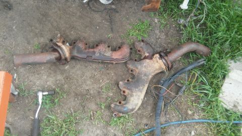 1979 Ford Bronco - Two old, rusty exhaust manifolds lie on the ground next to some grass and wiring.