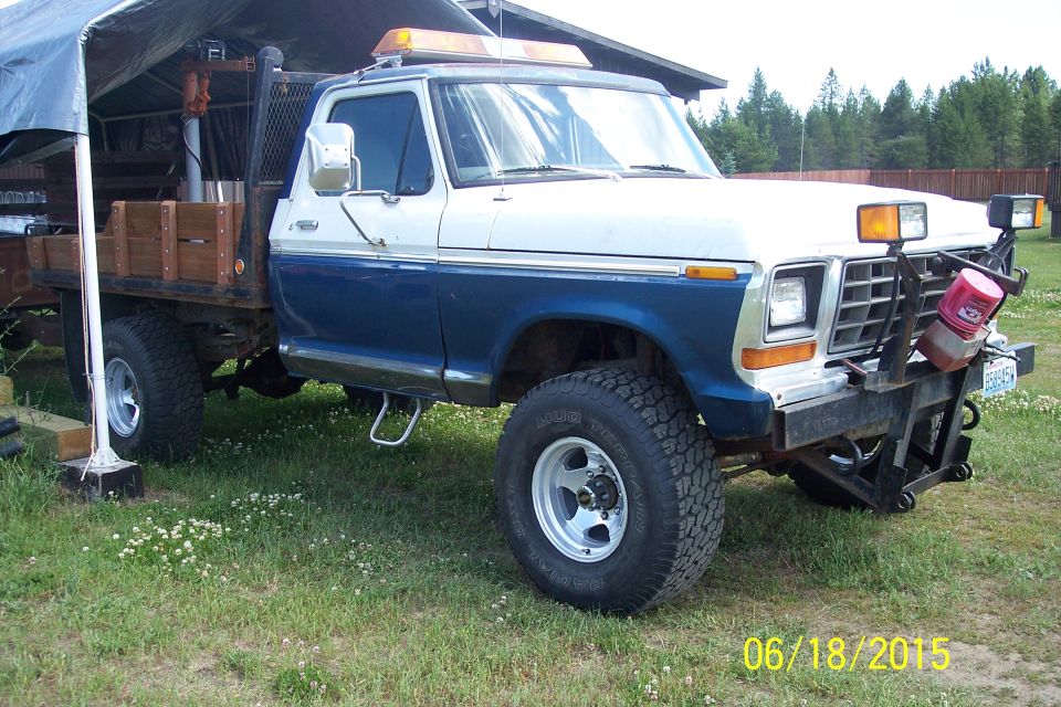 Ford F-Series pickup with flatbed, dual rear wheels, and a plow mount. It has a two-tone blue and white paint scheme and an amber light bar on the roof