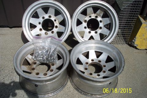 1989 Ford Bronco - A set of four aluminum alloy wheels are displayed on a concrete surface, with a bag of lug nuts nearby.