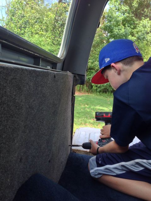 1979 Ford Bronco - A person works on the interior of a vehicle, using a drill on the carpeted panel.