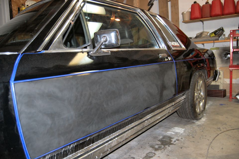 Black 1985 Mercury Grand Marquis side view, showing a blue pinstripe and a matte black panel on the door