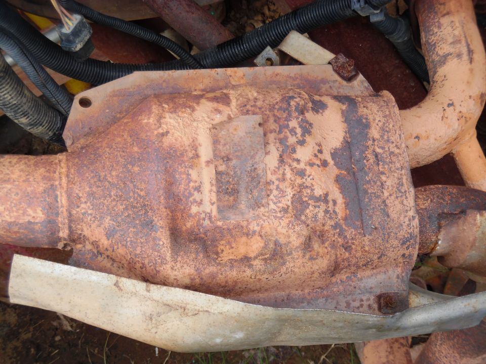 Rusted catalytic converter with bolted heat shield removed and lying among old exhaust pipes