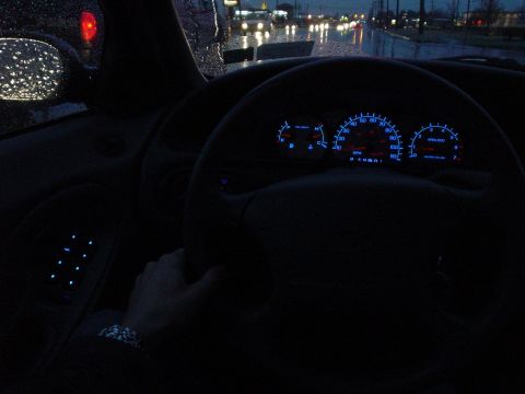 1999 Ford Taurus - Driver's-eye interior shot at dusk in the rain, showing a blue-backlit three-pod instrument cluster with a 120-mph speedometer, tachometer…