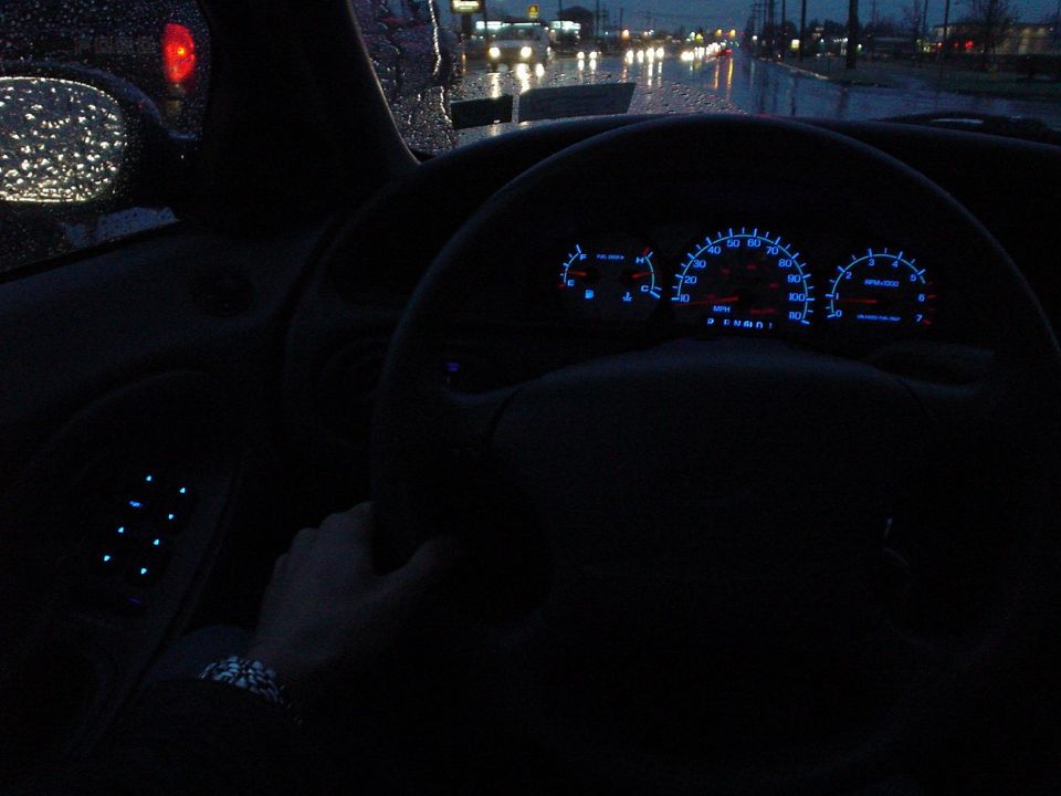 Blue-lit instrument cluster and steering wheel seen from the driver's seat at dusk in rain